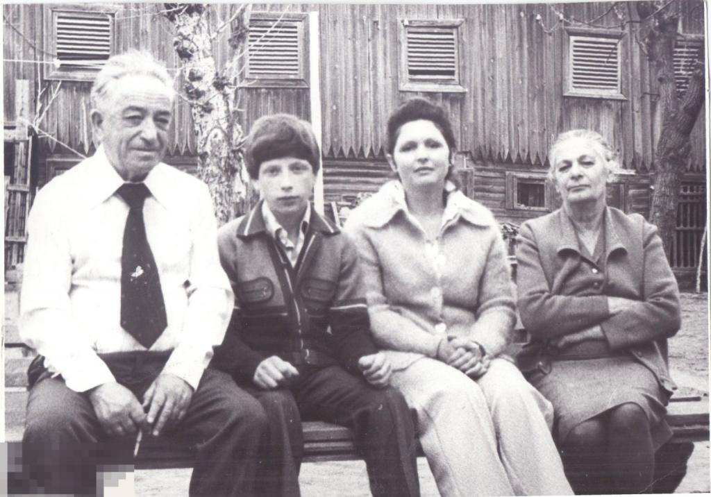 Семья сидит на скамейке на фоне деревянной постройки. 1980-е гг.